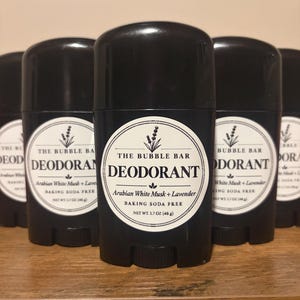 May include: A black plastic deodorant stick container with a white circular label. The label features the text THE BUBBLE BAR DEODORANT, Arabian White Musk + Lavender, BAKING SODA FREE, and NET WT. 1.7 OZ (48 g) along with a small lavender illustration.