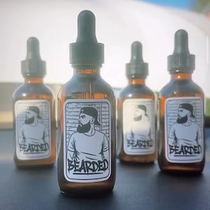 Puede incluir: Cuatro botellas de vidrio ámbar con cuentagotas negros. Cada botella tiene una etiqueta blanca con una ilustración en blanco y negro de un hombre con barba y la palabra "BEARDED". Las botellas están dispuestas en fila.