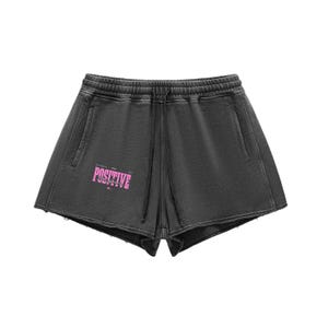 Può includere: Pantaloncini grigio scuro con vita con coulisse e tasche. I pantaloncini presentano la scritta "POSITIVE" in rosa sul davanti. I pantaloncini hanno un aspetto usurato.