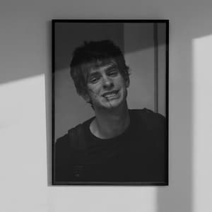 May include: Black and white framed portrait of a smiling person with short hair. The person is wearing a dark shirt and has a small scar on their face. The portrait is hanging on a white wall.