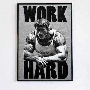 Puede incluir: Un póster enmarcado con una imagen en escala de grises de un culturista con una camiseta sin mangas. Las palabras "WORK HARD" están impresas en letras negras grandes y en negrita encima y debajo de la imagen. El póster está sobre un fondo gris claro.