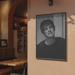 May include: Black and white framed portrait of a smiling person with short hair and a dark shirt. The portrait is hanging on a peach-colored wall outside a cafe. The frame is black.