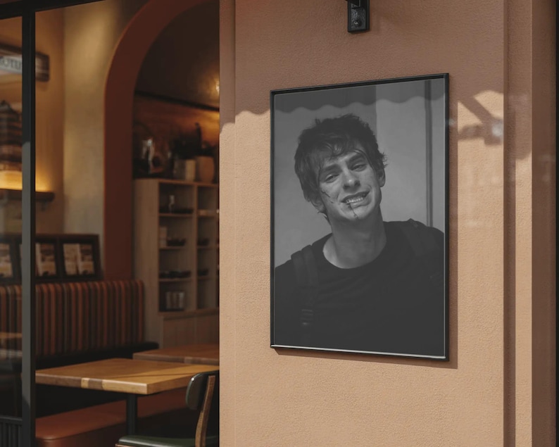 May include: Black and white framed portrait of a smiling person with short hair and a dark shirt. The portrait is hanging on a peach-colored wall outside a cafe. The frame is black.