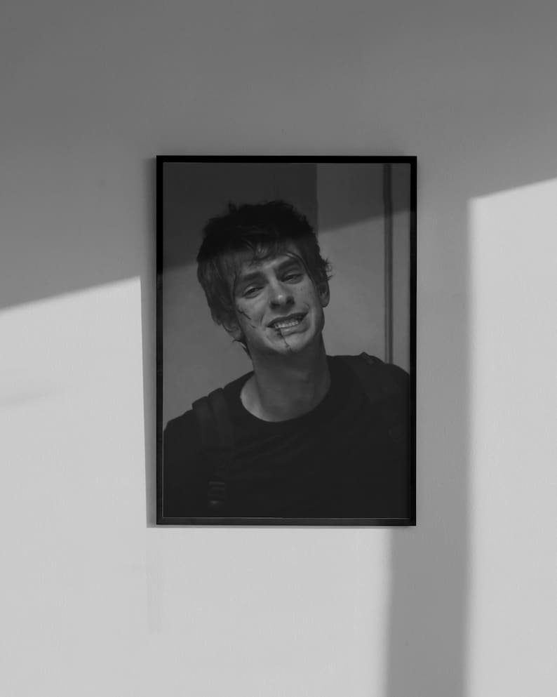 May include: Black and white framed portrait of a smiling person with short hair. The person is wearing a dark shirt and has a small scar on their face. The portrait is hanging on a white wall.