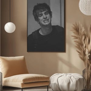 May include: Black and white framed portrait of a person with a scar on their face. The person is wearing a black shirt. The portrait is hanging on a tan wall. A beige chair and ottoman are in the foreground.