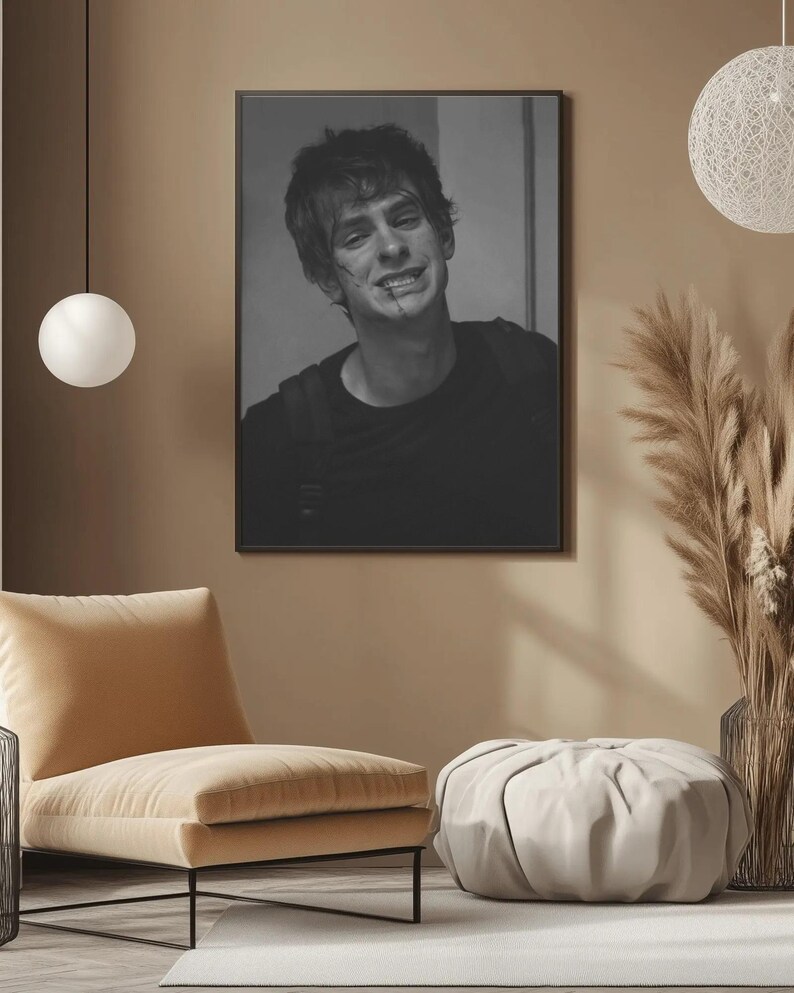 May include: Black and white framed portrait of a person with a scar on their face. The person is wearing a black shirt. The portrait is hanging on a tan wall. A beige chair and ottoman are in the foreground.