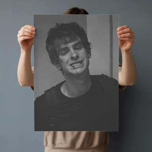 May include: Black and white print of a smiling person with a backpack. The person has short, messy hair and a few marks on their face. The print is held up by a person in a tan shirt.