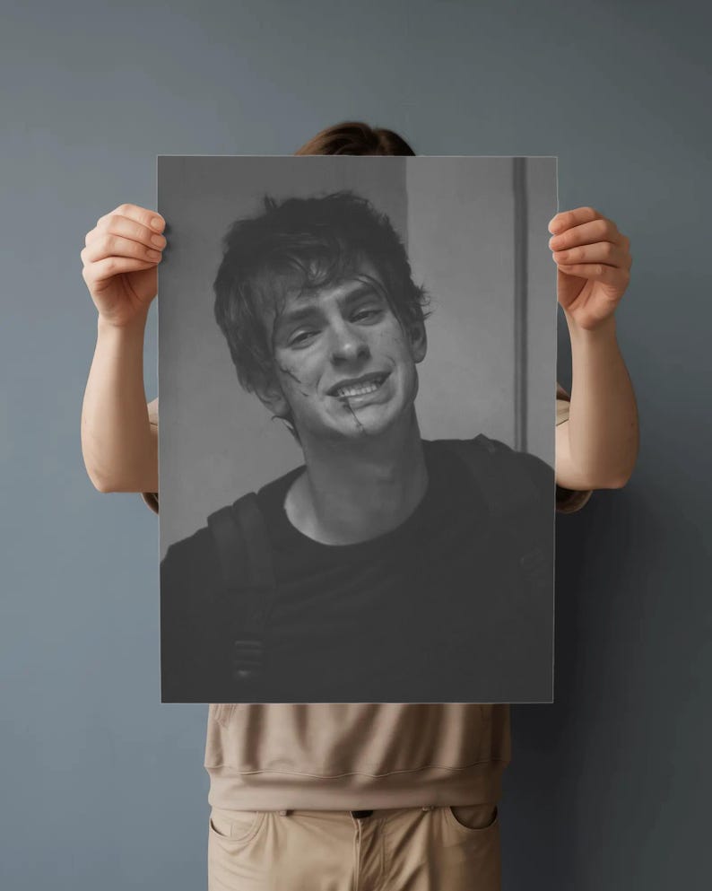 May include: Black and white print of a smiling person with a backpack. The person has short, messy hair and a few marks on their face. The print is held up by a person in a tan shirt.