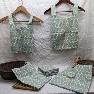 Puede incluir: Conjunto de cinco bolsas de la compra reutilizables con un patrón de hojas verdes y grises. Las bolsas están hechas de tela y tienen un diseño sencillo. Están colgadas de perchas de madera y dispuestas sobre una superficie blanca.