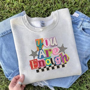 May include: A light gray sweatshirt with the embroidered phrase "YOU ARE ENOUGH" in colorful, block-letter style. The design includes checkered stars and a checkered border. The sweatshirt is paired with blue denim jeans.