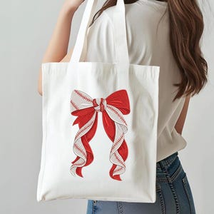 May include: White tote bag featuring a large red bow with white and red baseball stitching details. The bow design is centered on the bag, creating a striking visual contrast against the white fabric. The bag has two white handles.