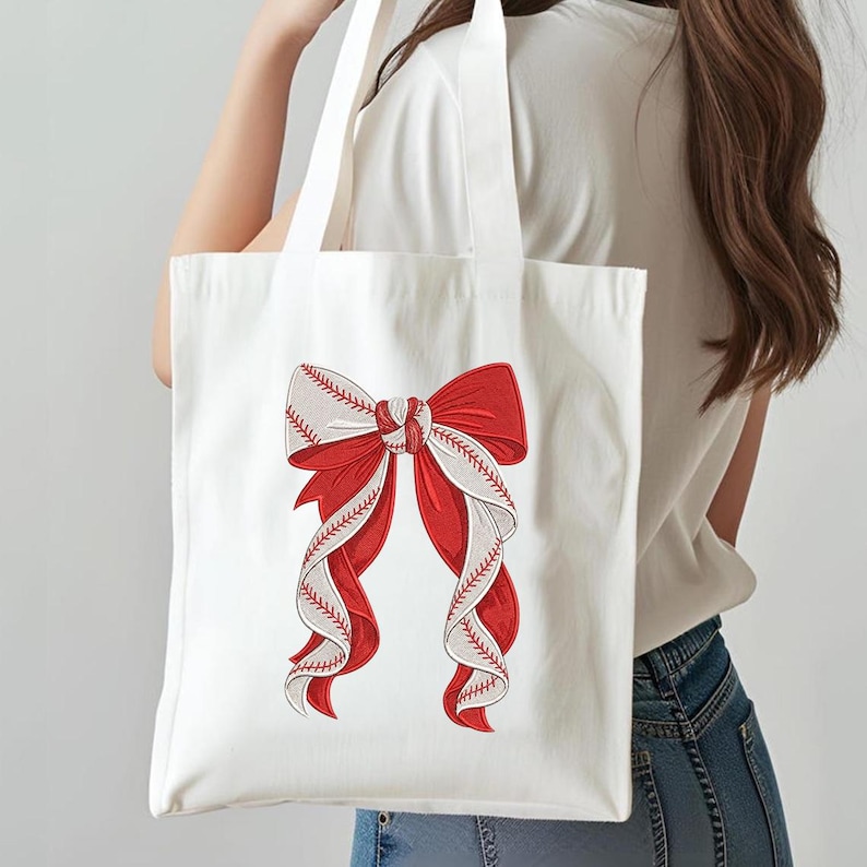 May include: White tote bag featuring a large red bow with white and red baseball stitching details. The bow design is centered on the bag, creating a striking visual contrast against the white fabric. The bag has two white handles.