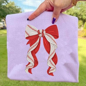 May include: A lavender tote bag featuring a baseball-themed bow design. The bow is a combination of red and white, with the white sections resembling baseballs, complete with red stitching. The bag is held up by a hand.