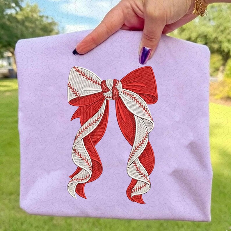 May include: A lavender tote bag featuring a baseball-themed bow design. The bow is a combination of red and white, with the white sections resembling baseballs, complete with red stitching. The bag is held up by a hand.