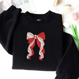 May include: Black sweatshirt featuring a red and white bow design. The bow is detailed with baseball stitching, creating a unique and sporty aesthetic. The sweatshirt has a crew neck and long sleeves, offering a casual and comfortable style.