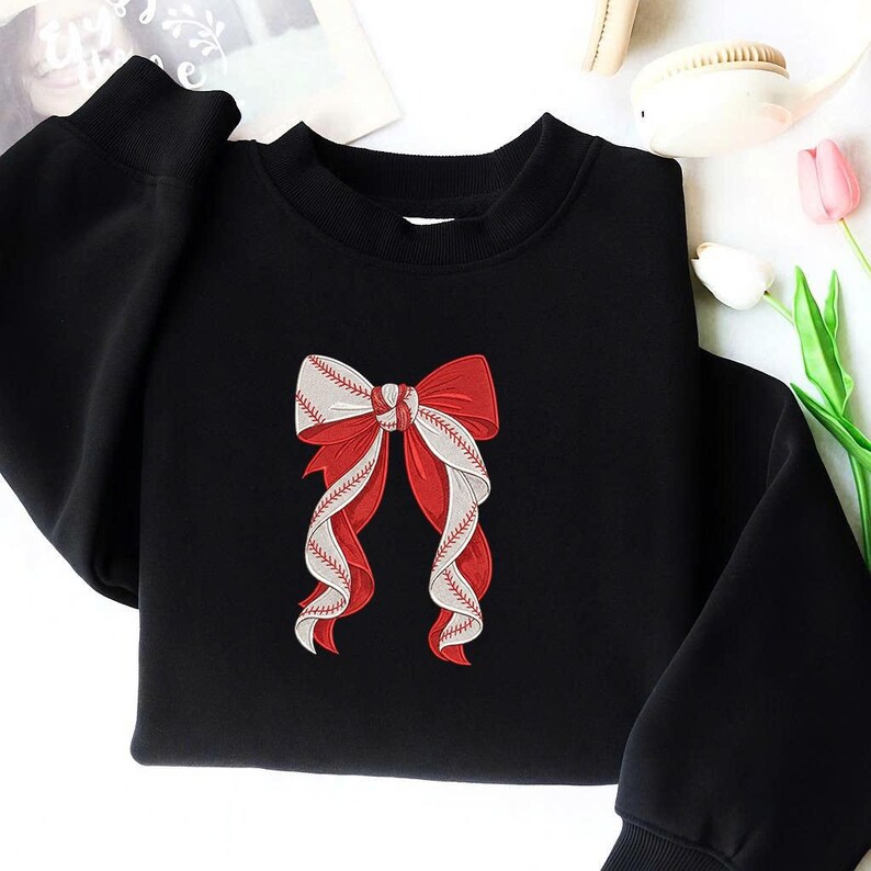 May include: Black sweatshirt featuring a red and white bow design. The bow is detailed with baseball stitching, creating a unique and sporty aesthetic. The sweatshirt has a crew neck and long sleeves, offering a casual and comfortable style.
