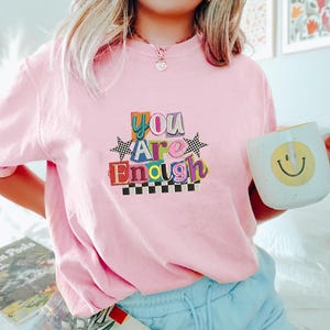 May include: A light pink t-shirt with the words "You Are Enough" in colorful, block letters. The shirt features a checkerboard design and star accents. A white mug with a yellow smiley face is held in the hand.