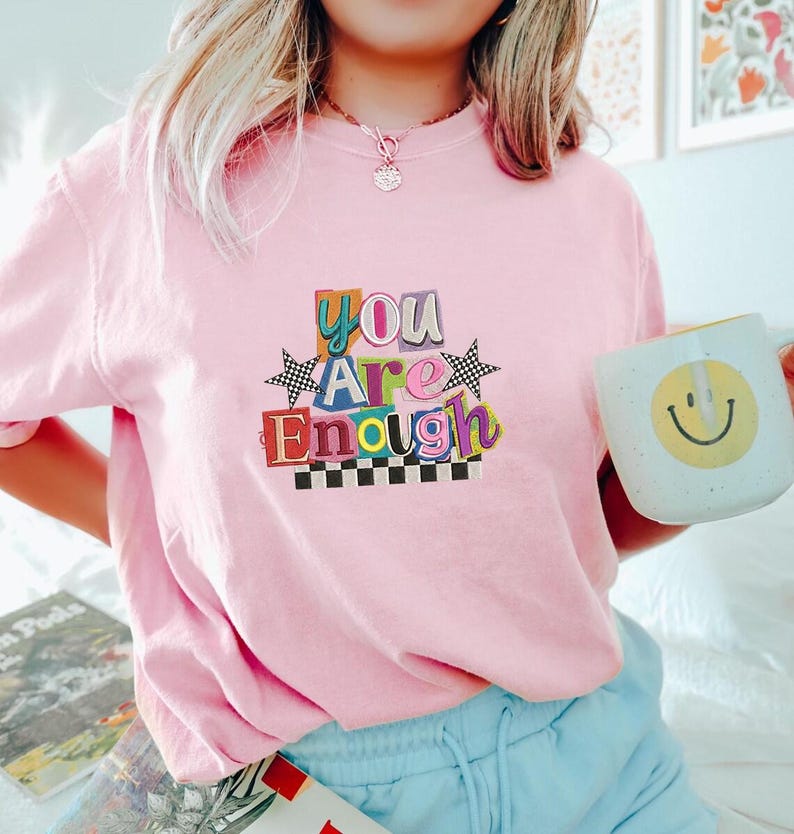 May include: A light pink t-shirt with the words "You Are Enough" in colorful, block letters. The shirt features a checkerboard design and star accents. A white mug with a yellow smiley face is held in the hand.