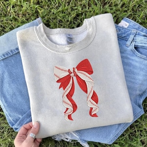 May include: A light gray sweatshirt featuring a large red and white bow design. The bow incorporates baseball stitching details, creating a unique sports-themed aesthetic. The sweatshirt is paired with blue denim jeans.