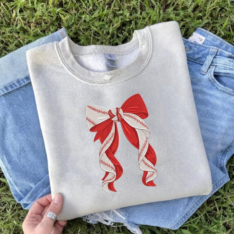 May include: A light gray sweatshirt featuring a large red and white bow design. The bow incorporates baseball stitching details, creating a unique sports-themed aesthetic. The sweatshirt is paired with blue denim jeans.