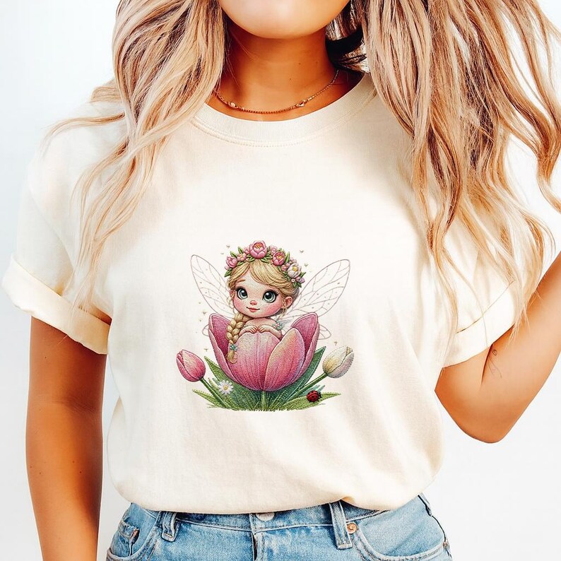 May include: A cream-coloured t-shirt featuring a graphic print of a small fairy with blonde hair and a flower crown, sitting inside a large pink tulip. The design includes green leaves, small white flowers, and a tiny red ladybug at the base.