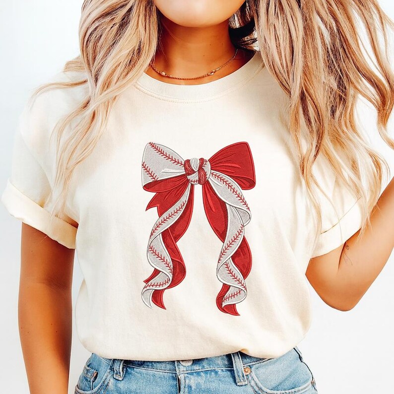 May include: Cream-colored t-shirt featuring a baseball-themed bow design. The bow has red and white sections, with baseball stitching details. The shirt's sleeves are rolled up, and the overall style is casual.