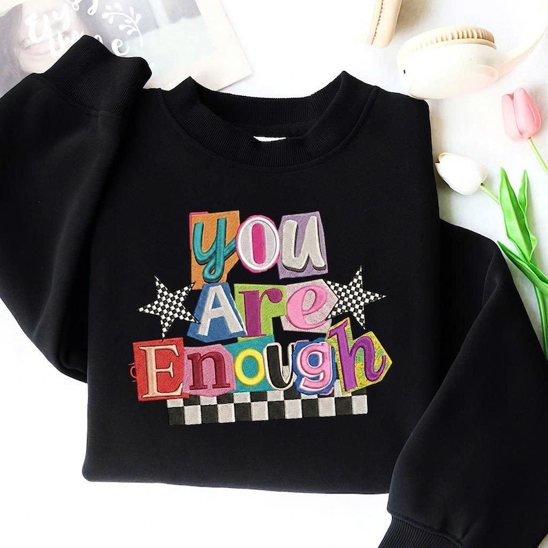 May include: Black sweatshirt with the embroidered text "You Are Enough" in colorful, block-letter style. The design includes a checkerboard border and star accents. The sweatshirt has a crew neck and long sleeves.
