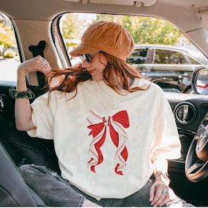 May include: Cream-colored t-shirt featuring a large red and white baseball-themed bow design. The bow has red ribbons and white ribbons with baseball stitching details. The shirt is worn by a person in a car.