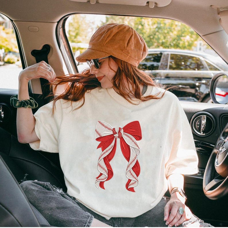 May include: Cream-colored t-shirt featuring a large red and white baseball-themed bow design. The bow has red ribbons and white ribbons with baseball stitching details. The shirt is worn by a person in a car.