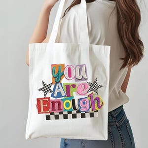 May include: White tote bag with the embroidered phrase "You Are Enough" in colorful, block letters. The design includes two black and white checkered stars and a checkered border at the bottom. The bag has two white handles.