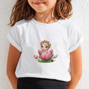 May include: A white short-sleeved t-shirt featuring a central graphic of a blonde fairy with a flower crown sitting inside a large pink tulip. The illustration includes green leaves, smaller tulip buds, and a tiny ladybug at the base.