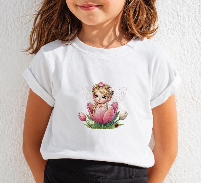 May include: A white short-sleeved t-shirt featuring a central graphic of a blonde fairy with a flower crown sitting inside a large pink tulip. The illustration includes green leaves, smaller tulip buds, and a tiny ladybug at the base.