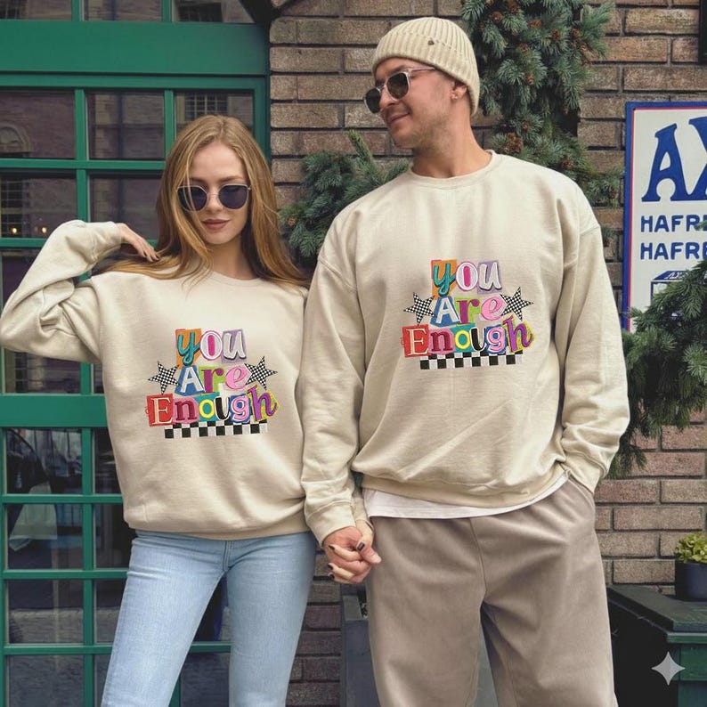 May include: Two people wearing matching beige sweatshirts with the text "You Are Enough" in colorful, retro-style lettering. The sweatshirts have a crew neck and long sleeves. One person is wearing blue jeans, and the other is wearing beige pants.