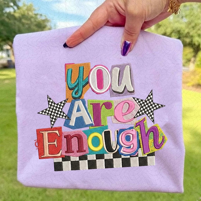 May include: A lavender tote bag with the embroidered words "YOU ARE ENOUGH" in colorful, block-letter style. The design includes two checkered stars and a checkered border at the bottom.