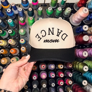 May include: A beige and black trucker hat with the words "DANCE mom" embroidered in black. The hat has a black brim and a mesh back. The background features a wall of colorful thread spools.