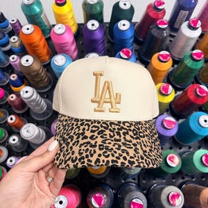 May include: A cream-coloured baseball cap embroidered with the letters "LA" in gold. The brim has a leopard print design. The background displays spools of colourful thread, indicating a craft or sewing theme.