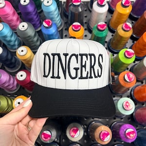 May include: A white baseball cap with black pinstripes and a black peak. The word "DINGERS" is embroidered in large, black letters. The background shows a variety of colourful thread spools, indicating a craft or sewing project.