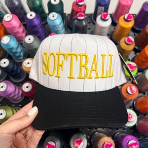 Op de afbeelding: Witte en zwarte softbalpet met het woord "SOFTBALL" in geel geborduurd. De pet heeft een zwarte klep en een witte kroon met dunne zwarte verticale strepen. De achtergrond toont spoelen met kleurrijk garen.