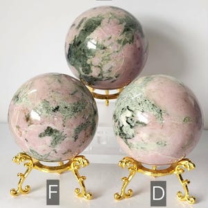 May include: Three polished spherical stones with a mottled pink and green pattern, each resting on a decorative gold-toned metal tripod stand. The spheres are labelled with the letters C, F, and D, indicating distinct individual items.