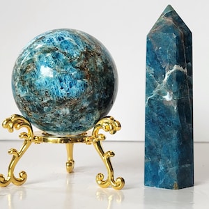 Op de afbeelding: Een gepolijste blauwe apatietbol rustend op een decoratieve goudkleurige metalen driepootstandaard, naast een hoge, veelzijdige blauwe apatiet kristaltoren. Beide mineralen vertonen diepblauwe tinten met natuurlijke bruine en witte insluitingen.