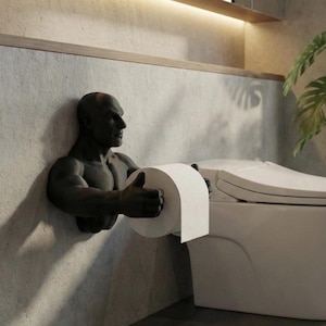 May include: A dark grey wall-mounted toilet roll holder sculpted as a muscular man holding a roll of white toilet paper. The holder is on a textured grey wall next to a white toilet.