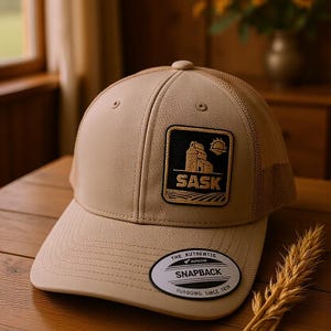 Könnte beinhalten: Beigefarbene Snapback-Kappe mit Mesh-Rückseite und gebogenem Schirm. Die Kappe hat einen quadratischen Aufnäher mit einer Grafik eines Gebäudes, einer Sonne und dem Wort "SASK". Sie hat auch ein Etikett mit der Aufschrift "THE AUTHENTIC SNAPBACK YUPOONG SINCE 1974".
