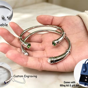 Op de afbeelding: Twee zilverkleurige, open te maken armbanden met schroefdoppen en groene en witte steenaccenten. Eén inzetstuk toont de armband open, een andere toont blauwe steenuiteinden met gravure, en een derde toont een blauwe geschenkdoos met vulset.