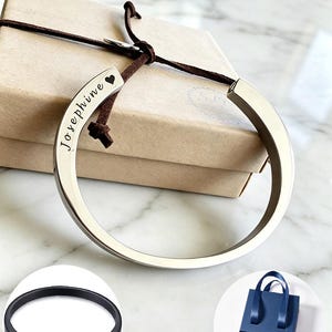 Könnte beinhalten: Ein silberfarbener Metallarmreif mit der Gravur Josephine und einem Herzsymbol, versehen mit einem braunen Lederband. Das Set enthält einen schwarzen, zu öffnenden Armreif, ein Befüllungsset und eine blaue Geschenkbox für das Schmuckstück.