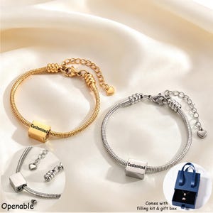 Könnte beinhalten: Zwei Schlangenketten-Armbänder, eines in Goldoptik und eines in Silberoptik, mit jeweils einem zentralen Würfelanhänger, der mit dem Wort Customized graviert ist. Beide Armbänder haben einen Karabinerverschluss, eine verstellbare Verlängerungskette mit Herzanhänger und sind zum Öffnen.