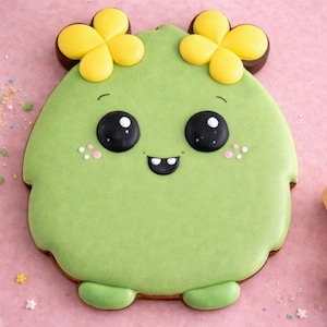 May include: A green, cartoon-like cookie with large black eyes, a small smile, and yellow flower-shaped accents. The cookie has a light green frosting and is decorated with small pink dots. The cookie is on a pink surface.