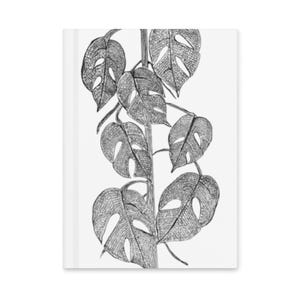 May include: A white notebook featuring a detailed black and white illustration of a Monstera plant. The drawing showcases the plant's distinctive split leaves and stem, creating a botanical theme.