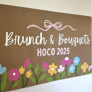 May include: A brown sign with the words "Brunch & Bouquets" in white script, and "HOCO 2025" in pink. A pink bow and colorful flowers in pink, yellow, blue, and white decorate the bottom.