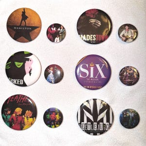 May include: A collection of round, colorful buttons featuring musical theater productions. The buttons display logos and imagery from shows like "Hamilton," "Wicked," "Six," and "Beetlejuice." The buttons vary in size and design, with vibrant colors and text.