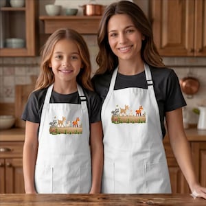 May include: Two white aprons with a woodland animal design. The aprons feature a raccoon, deer, rabbit, and fox on a log with mushrooms and grass. The aprons are worn by a woman and a child.